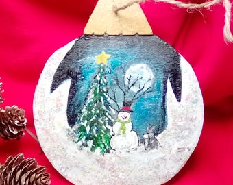 May include: A hand-painted wooden ornament shaped like a Christmas tree with a snowman and a pine tree in the snow. The ornament is decorated with glitter and has a blue, white, and green color scheme.