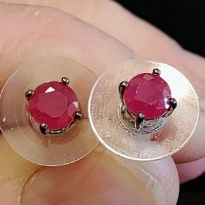 May include: A pair of stud earrings featuring round, faceted, deep red gemstones. Each gem is set in a dark metal prong setting and mounted on a clear, circular backing. The earrings are shown close up.