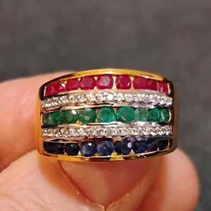 May include: A gold band ring with multiple rows of gemstones. The top row features red rubies, followed by a row of clear diamonds, then a row of green emeralds, another row of diamonds, and a final row of blue sapphires.