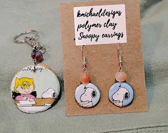 May include: A keychain and earrings set featuring Snoopy and other Peanuts characters. The keychain has a round charm with a cartoon image, a silver keyring, and a purple bead. The earrings have Snoopy images and peach-colored beads.