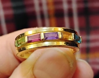 May include: A gold band ring with five different colored gemstones. The gemstones are green, pink, purple, orange, and blue.