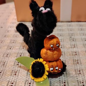 May include: A black cat figurine with a pink nose sits next to a stack of three orange pumpkin decorations. A yellow and black sunflower with green leaves is in front of the pumpkins. The objects are on a patterned surface.