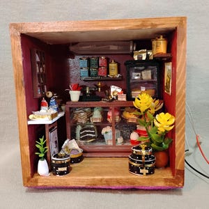 May include: Miniature bakery diorama in a wooden box. The interior features a display case with cakes and pastries, shelves with tea tins, and decorative flowers. The scene is lit with small lights and includes a small table with milk bottles.
