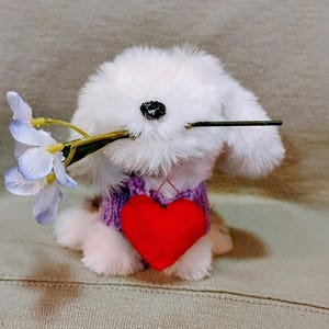 May include: A fluffy white stuffed bunny wearing a purple sweater and a red heart. The bunny holds a sprig of light purple flowers in its mouth. The toy is sitting on a neutral-colored surface.