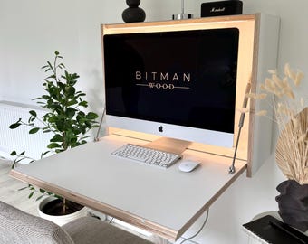 May include: A wall-mounted white desk with a light wood edge, featuring a fold-down work surface supported by metal struts. The desk includes integrated interior lighting and holds a computer monitor displaying the text BITMAN WOOD.