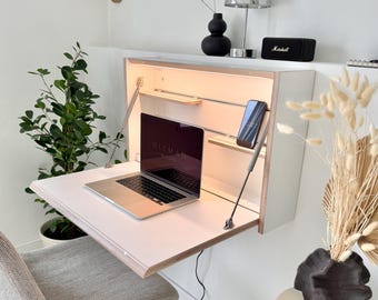 May include: A wall-mounted white fold-down desk with a light wood edge, featuring an integrated internal light, a silver laptop, and a smartphone. The desk is supported by two metal struts and is positioned above a grey upholstered chair.