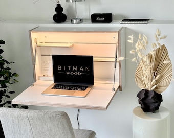 May include: A wall-mounted white desk with a fold-down work surface and integrated interior lighting. The desk features a light wood edge finish and contains a laptop with the text BITMAN WOOD on the screen. A grey upholstered chair sits in front.