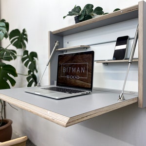 Könnte beinhalten: Ein grauer, an der Wand montierter Klapptisch mit einem aufgeschlagenen Laptop auf der Oberfläche. Der Tisch hat ein Regal darüber mit einem Smartphone auf dem Regal. Der Tisch ist aus Sperrholz und hat eine graue Oberfläche. Der Laptop-Bildschirm zeigt den Text "BITMAN WOOD" an.