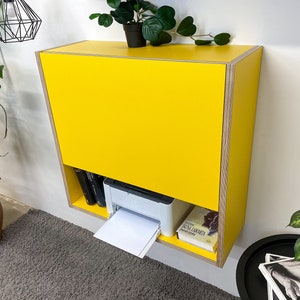 May include: A wall-mounted yellow cabinet with a plywood edge finish. The cabinet features a large solid yellow front panel and an open lower shelf containing a white printer, a black book, and a book with a light cover titled Juna Larmeta.