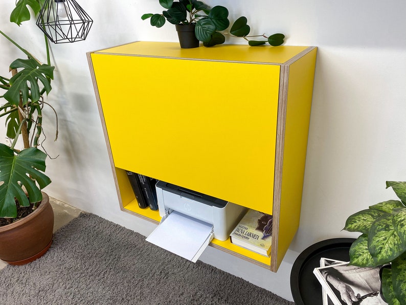 May include: A wall-mounted yellow cabinet with a plywood edge finish. The cabinet features a large solid yellow front panel and an open lower shelf containing a white printer, a black book, and a book with a light cover titled Juna Larmeta.