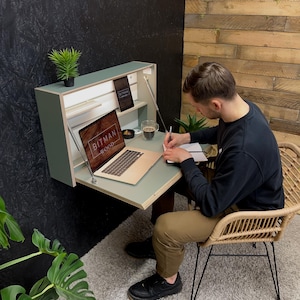 Könnte beinhalten: Ein grüner, an der Wand montierter Klapptisch mit Laptop, Notizbuch und Stift. Der Tisch ist geöffnet und eine Person sitzt darauf in einem Korbstuhl.