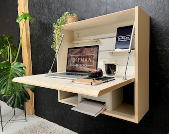 May include: A light-colored, wall-mounted desk with a laptop, tablet, and printer. The laptop and tablet display the "BITMAN WOOD" logo. The desk is open, revealing a workspace with a coffee cup and a small bowl.