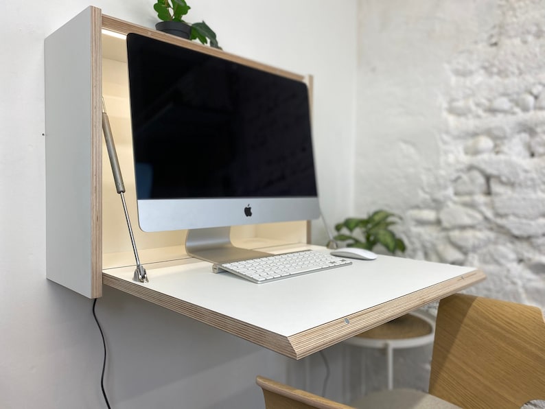 Puede incluir: Un escritorio plegable montado en la pared con superficie blanca y bordes de madera contrachapada a la vista. El escritorio incluye luz integrada, un monitor de computadora plateado, un teclado inal&aacute;mbrico blanco y un rat&oacute;n blanco sobre la mesa.
