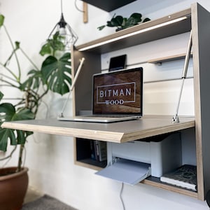 Può includere: Una scrivania a parete pieghevole in compensato grigio scuro con bordi a vista. La scrivania presenta un piano di lavoro ribaltabile sostenuto da montanti in metallo, luce LED integrata, ripiani interni e un portatile con il testo BITMAN WOOD.