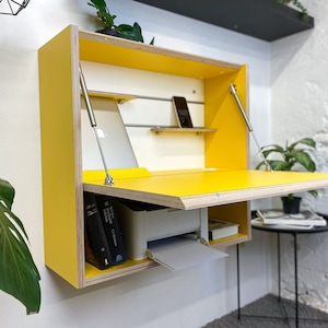 May include: A wall-mounted yellow desk with a fold-down work surface supported by two metal struts. The interior features a white back panel with a horizontal metal rail, a small shelf holding a smartphone, and a lower compartment containing a printer.