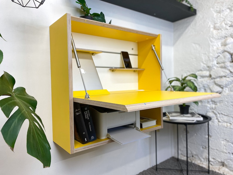 May include: A wall-mounted yellow desk with a fold-down work surface supported by two metal struts. The interior features a white back panel with a horizontal metal rail, a small shelf holding a smartphone, and a lower compartment containing a printer.