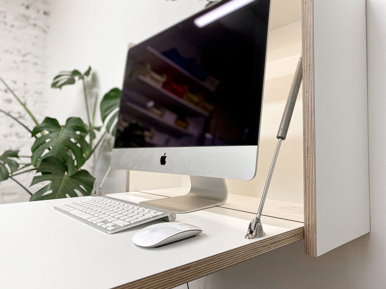 Puede incluir: Un escritorio blanco montado en la pared con borde de madera contrachapada, que cuenta con una superficie abatible sostenida por dos pistones de gas met&aacute;licos. Un Apple iMac, un teclado inal&aacute;mbrico blanco y un rat&oacute;n blanco est&aacute;n sobre la mesa.