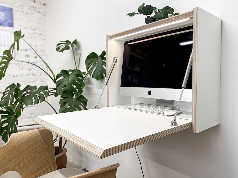 Puede incluir: Un escritorio blanco montado en la pared con una superficie de trabajo abatible, con bordes de madera contrachapada expuestos y dos soportes met&aacute;licos. El interior incluye una tira de luz integrada, un monitor, un teclado y un rat&oacute;n.