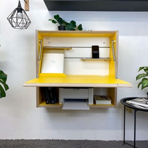 May include: A wall-mounted, fold-down desk featuring a bright yellow interior and light wood grain exterior. The unit includes a drop-down work surface, internal shelving, metal support struts, and a white printer housed in the lower compartment.