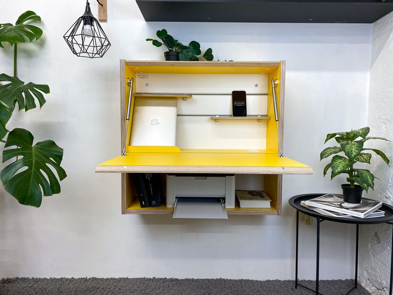 May include: A wall-mounted, fold-down desk featuring a bright yellow interior and light wood grain exterior. The unit includes a drop-down work surface, internal shelving, metal support struts, and a white printer housed in the lower compartment.
