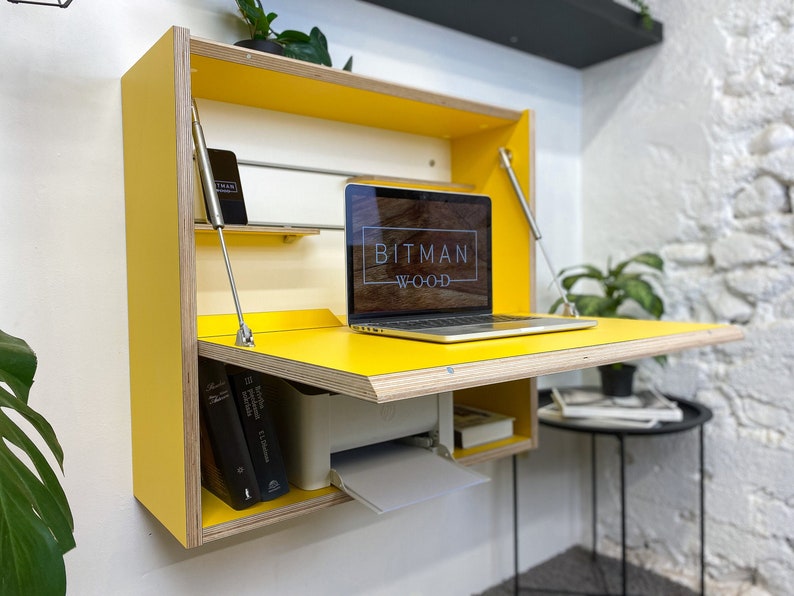 May include: A wall-mounted yellow desk with a fold-down work surface, featuring exposed plywood edges, metal support struts, and an internal shelf. A laptop with the text BITMAN WOOD on the screen sits on the desk, with books and a printer stored inside.