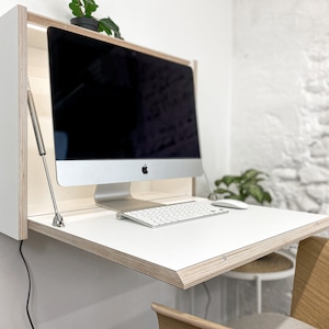 May include: A wall-mounted fold-down desk featuring a white surface with exposed plywood edges. The unit includes a metal support strut on each side and holds a silver all-in-one computer, a white wireless keyboard, and a white computer mouse.