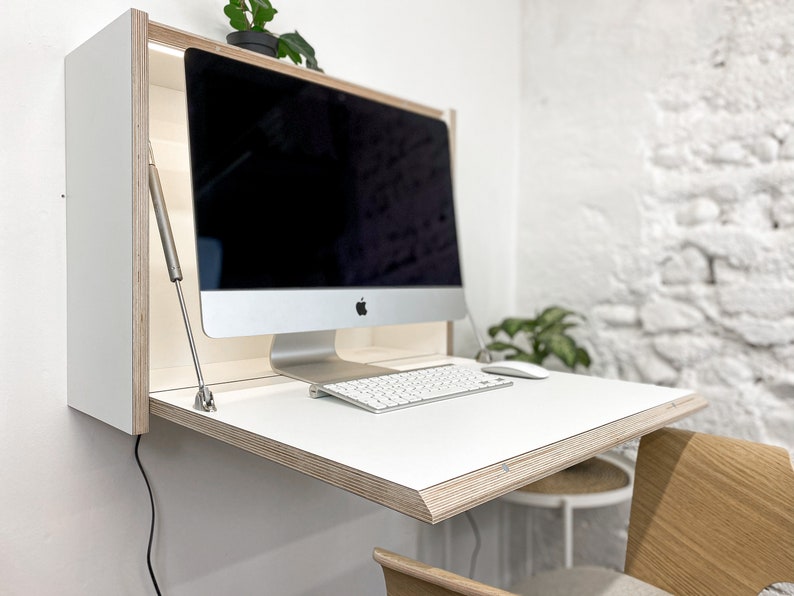 May include: A wall-mounted fold-down desk featuring a white surface with exposed plywood edges. The unit includes a metal support strut on each side and holds a silver all-in-one computer, a white wireless keyboard, and a white computer mouse.