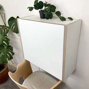 Puede incluir: Un armario rectangular montado en la pared con un panel frontal blanco liso y bordes de madera contrachapada clara expuestos. El armario se sit&uacute;a sobre una silla de madera con asiento tapizado en gris claro, creando un &aacute;rea de trabajo compacta.