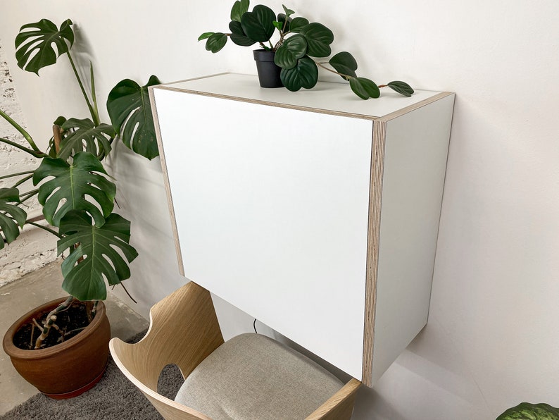 Puede incluir: Un armario rectangular montado en la pared con un panel frontal blanco liso y bordes de madera contrachapada clara expuestos. El armario se sit&uacute;a sobre una silla de madera con asiento tapizado en gris claro, creando un &aacute;rea de trabajo compacta.