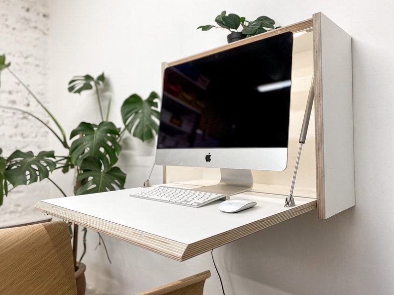 May include: A wall-mounted fold-down desk featuring a white surface with exposed plywood edges. The desk is equipped with two metal support struts on the sides and holds a silver all-in-one computer, a white wireless keyboard, and a white mouse.