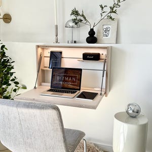 May include: A fold-down wall desk with a laptop displaying "BITMAN WOOD" text, a tablet, and a small speaker. The desk is open, revealing a shelf with a tablet and a small speaker. A grey chair and a white side table with a disco ball are in the foreground.