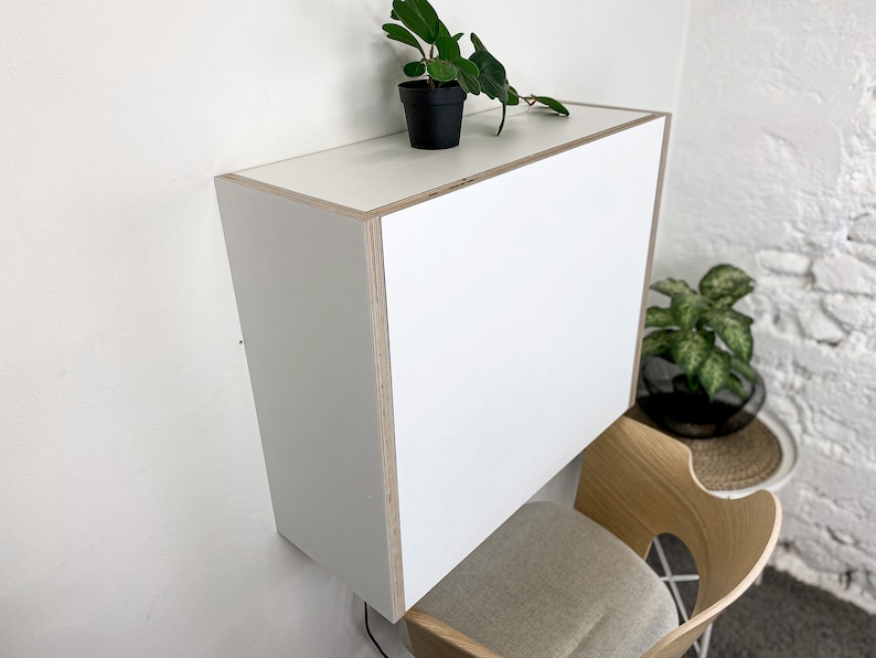 Puede incluir: Un escritorio blanco montado en la pared con forma rectangular y bordes de madera contrachapada de color claro expuestos. El escritorio cuenta con una superficie superior plana y un panel frontal liso, ofreciendo una soluci&oacute;n funcional.