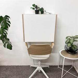 May include: A wall-mounted white desk with light wood side panels positioned above a chair with a curved light wood backrest, a light gray upholstered seat, and a white five-point base with casters, creating a compact workspace setup.