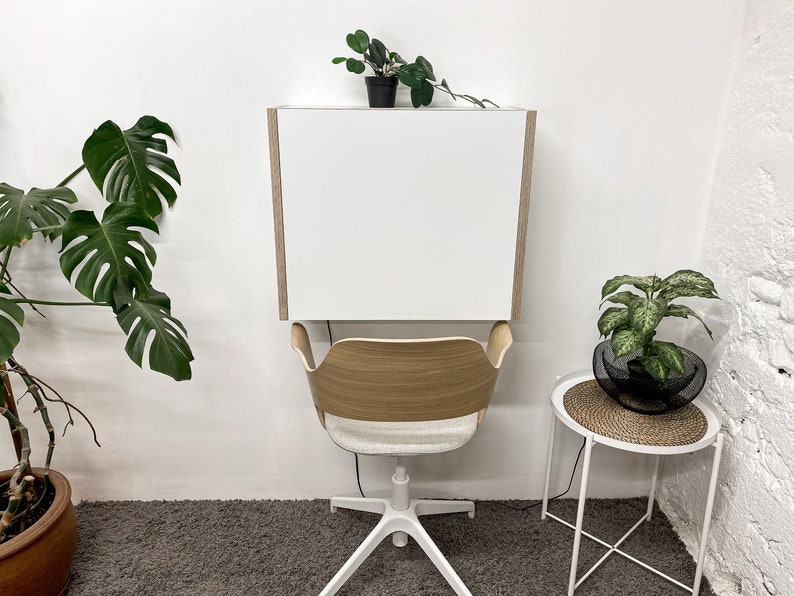 May include: A wall-mounted white desk with light wood side panels positioned above a chair with a curved light wood backrest, a light gray upholstered seat, and a white five-point base with casters, creating a compact workspace setup.