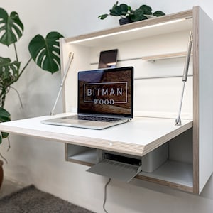 May include: A white wall-mounted fold-down desk with a laptop computer and a printer. The desk is made of plywood and has a light wood grain finish. The laptop screen displays the text "Bitman Wood".