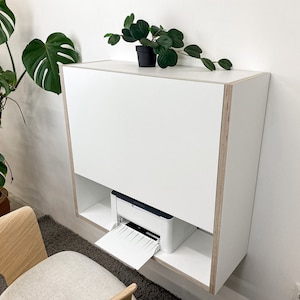 Peut inclure: Un bureau mural blanc avec un panneau avant rabattable et des bords en contreplaqué apparents. L'unité dispose d'une étagère inférieure ouverte contenant une imprimante blanche avec un bac à papier étendu. Le bureau a un design rectangulaire.