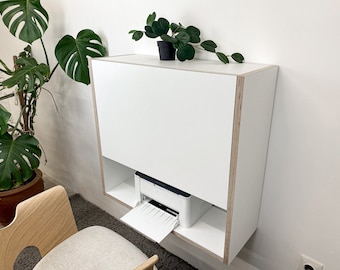 May include: A wall-mounted white desk with a fold-down front panel and exposed plywood edges. The unit features an open lower shelf containing a white printer with an extended paper tray. The desk has a clean, rectangular design with a flat top surface.