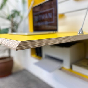 May include: A wall-mounted yellow desk with a plywood edge, featuring a fold-down work surface supported by metal struts. The desk interior contains a white printer and a book, while a laptop sits on the top surface with a visible logo.
