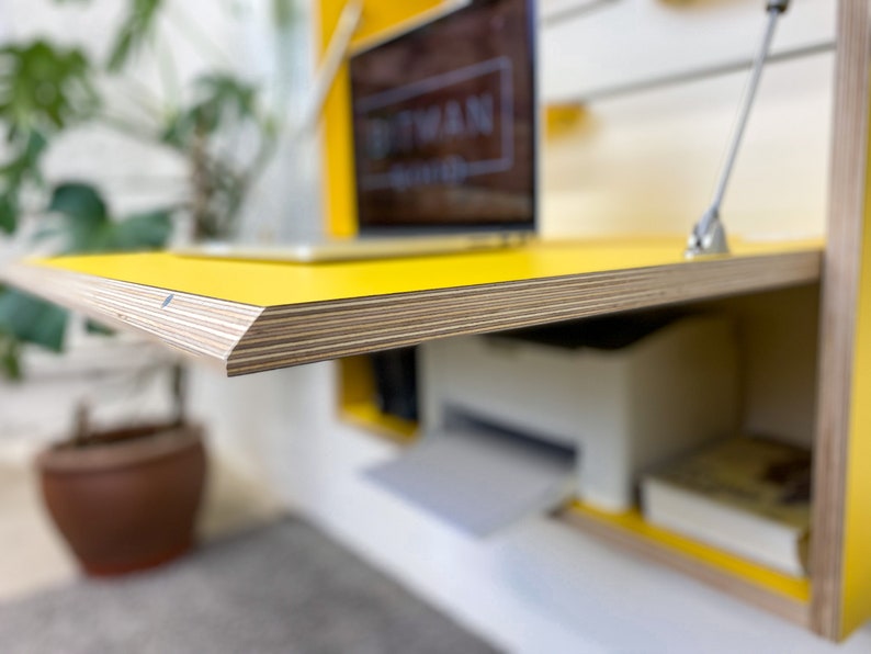 May include: A wall-mounted yellow desk with a plywood edge, featuring a fold-down work surface supported by metal struts. The desk interior contains a white printer and a book, while a laptop sits on the top surface with a visible logo.