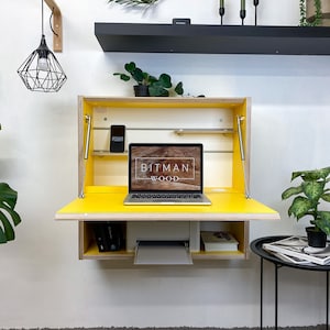May include: A wall-mounted wooden desk with a bright yellow fold-down work surface and interior. The unit features a laptop displaying the text BITMAN WOOD, a white printer on the lower shelf, and metal support struts on both sides of the frame.