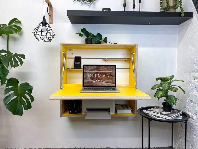 May include: A wall-mounted wooden desk with a bright yellow fold-down work surface and interior. The unit features a laptop displaying the text BITMAN WOOD, a white printer on the lower shelf, and metal support struts on both sides of the frame.