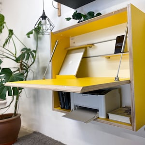 May include: A wall-mounted yellow desk with a fold-down work surface supported by two metal struts. The interior features a light-colored back panel with a horizontal shelf, a laptop, a printer, and a book stored within the wooden frame.