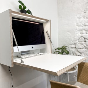 May include: A wall-mounted white desk with a fold-down work surface and exposed plywood edges. The unit features two metal support arms on the sides, an integrated light strip at the top, and a computer monitor with a keyboard placed inside.