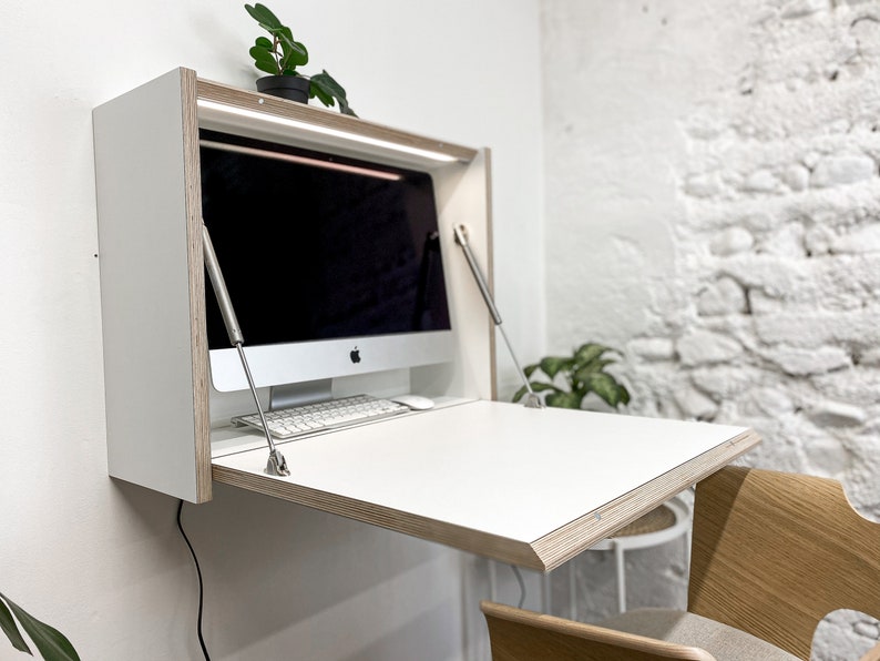 May include: A wall-mounted white desk with a fold-down work surface and exposed plywood edges. The unit features two metal support arms on the sides, an integrated light strip at the top, and a computer monitor with a keyboard placed inside.