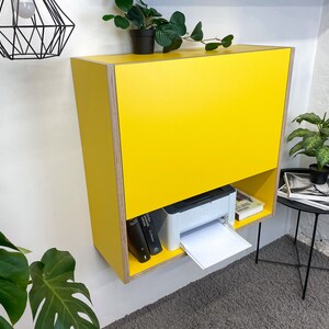 May include: A wall-mounted yellow cabinet with a flat front panel and an open lower shelf. The cabinet features exposed plywood edges and contains a white printer and two books on the bottom shelf. The unit is mounted on a plain white wall.