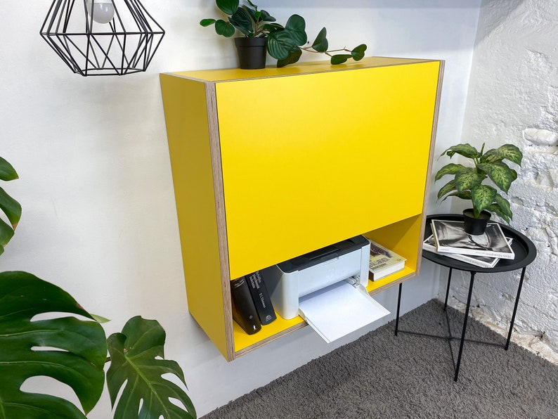 May include: A wall-mounted yellow cabinet with a flat front panel and an open lower shelf. The cabinet features exposed plywood edges and contains a white printer and two books on the bottom shelf. The unit is mounted on a plain white wall.