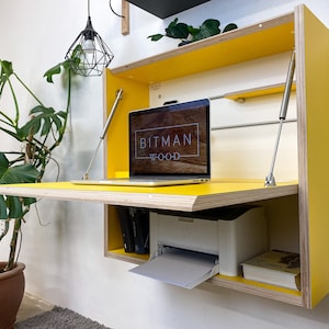May include: A wall-mounted, yellow desk with a laptop displaying the "BITMAN WOOD" logo. The desk is open, revealing a printer, books, and a shelf. The desk is supported by silver gas struts. A potted plant and a hanging light fixture are also visible.