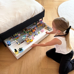 May include: A white under-bed storage drawer pulled out from beneath a black bed frame, containing a collection of colorful plastic building block sets, including small figures, vehicles, and miniature potted plant models arranged on a gray base.