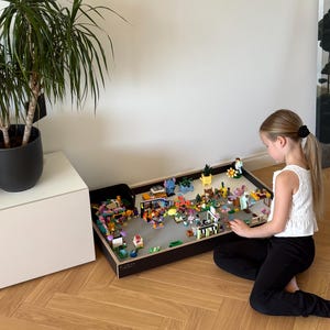 May include: A rectangular black wooden building block play table with a grey baseplate surface, featuring various colourful plastic construction toy figures, small structures, and accessories arranged across the flat play area for building activities.