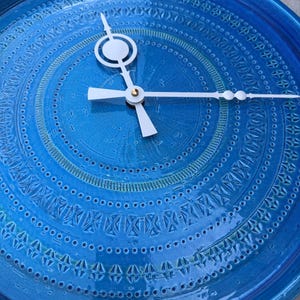 May include: A circular wall clock featuring a vibrant blue glazed ceramic face with intricate, embossed geometric patterns and concentric circles. The clock has three white hands, including a unique hour hand with a circular cutout design.